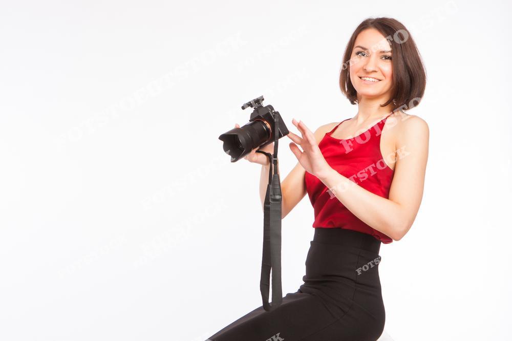 На фотографии изображена улыбающаяся женщина-фотограф с профессиональной камерой в руках. Она одета в яркий красный топ и черную юбку, что придает образу стильный и уверенный вид. Белый фон подчеркивает чистоту и профессионализм сцены, создавая акцент на творческой деятельности фотографа.